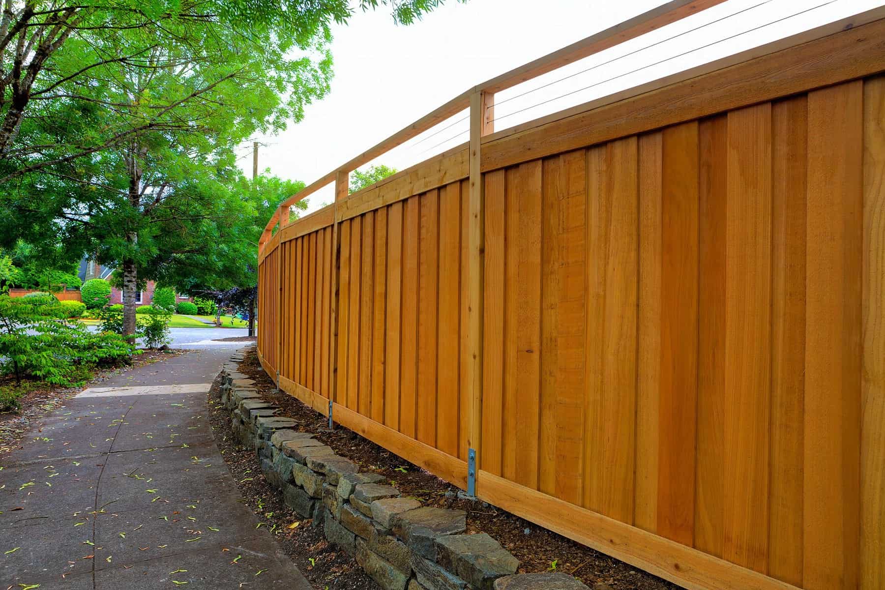 Cedar Stained Privacy Fence Golden-brown cedar-stained vertical board fence with white top rail and stone base in residential setting.